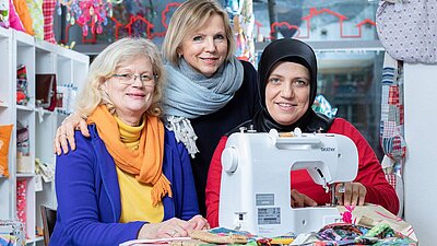 Drei Frauen sitzen und stehen vor einer Nähmaschine.