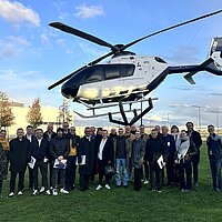 Eine Gruppe von Menschen steht vor einem in der Luft schwebenden Helikopter.