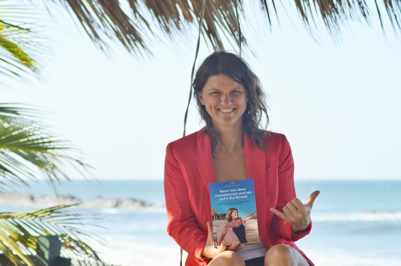 Fotografie einer Frau in einem roten Blazer die ein Buch in der Hand halt und eine Surfgeste mit der anderen Hand macht, sitzend am Strand. - copyright: Maike Simonis - Ersteller: Maike Simonis