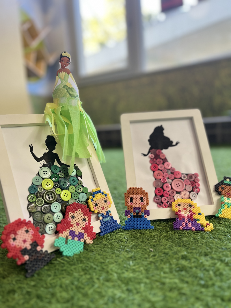 Bastelarrangement mit Disney-Prinzessinnen: Zwei Bilderrahmen mit Silhouettenkleidern aus Knöpfen (grün und rosa), davor kleine Figuren aus Bügelperlen und eine Tiana-Figur mit grünem Kleid, alles auf einer grasähnlichen Unterlage - copyright: Katrin Mumm - Ersteller: Katrin Mumm